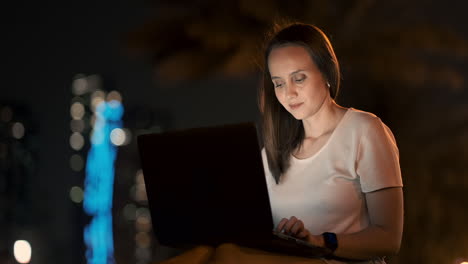 Young-beautiful-girl-designer-working-remotely-on-a-laptop-on-the-street-in-the-summer-under-palm-trees-at-night-in-the-city-Park.-United-Arab-Emirates-European-woman-at-night-in-Dubai